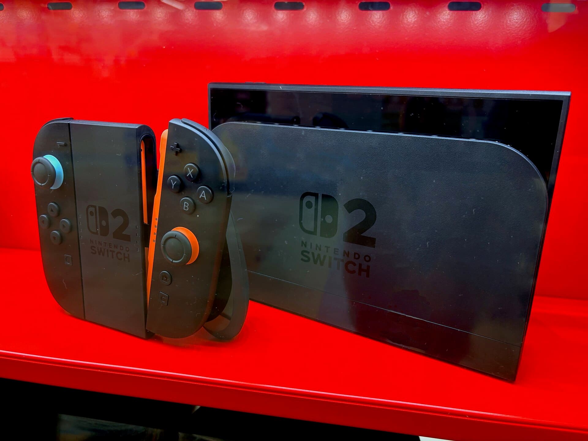 Nintendo Switch 2 console shown from behind with its kickstand, next to a pair of detached Joy-Con 2 controllers, on a red retail shelf