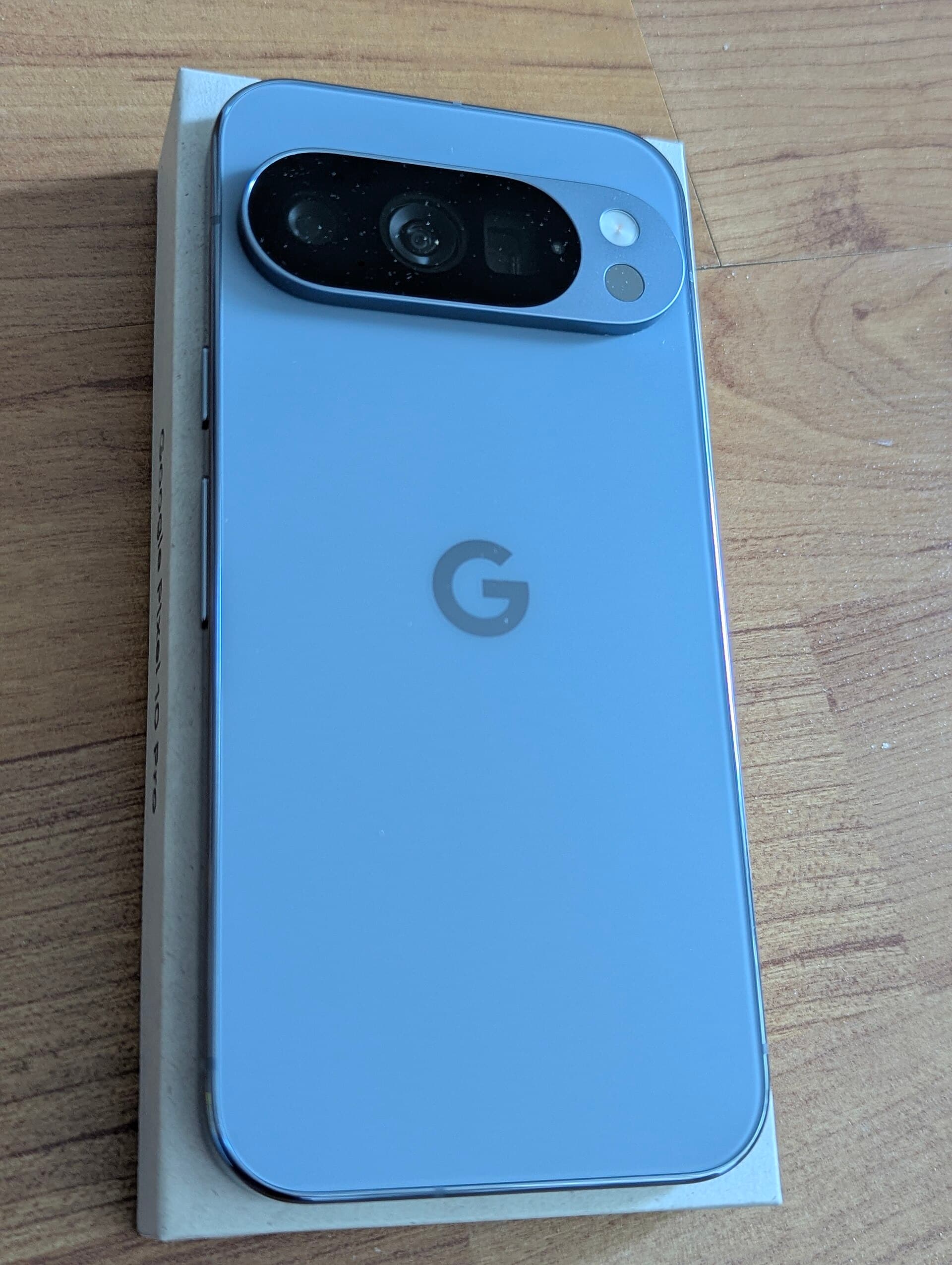Rear of a Google Pixel 10 Pro in Moonstone, showing the horizontal camera bar and the Google G logo, resting on its retail box